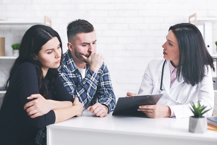 A female doctor in a white coat speaking with a worried couple sitting across her desk in a medical office.