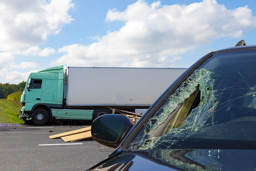 A shattered car windshield in the foreground with a green semi-truck involved in an accident in the background.