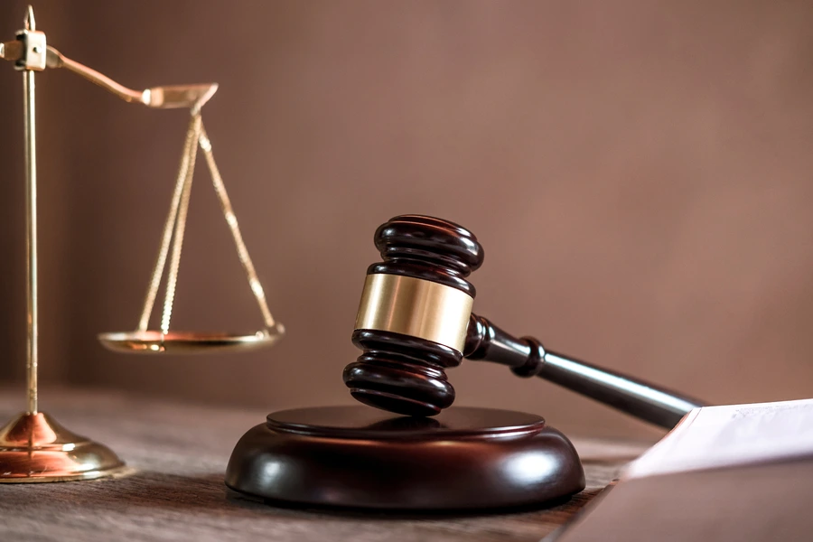 A wooden judge's gavel resting on a sounding block alongside a brass scale of justice and an open law book.