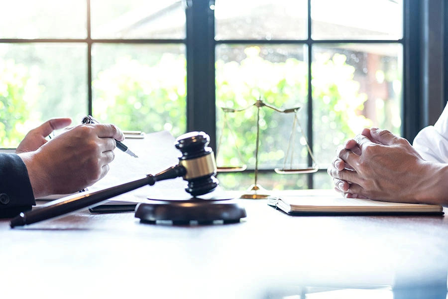 A legal consultation featuring a lawyer taking notes and a client with clasped hands, with a gavel and scale of justice on the table.