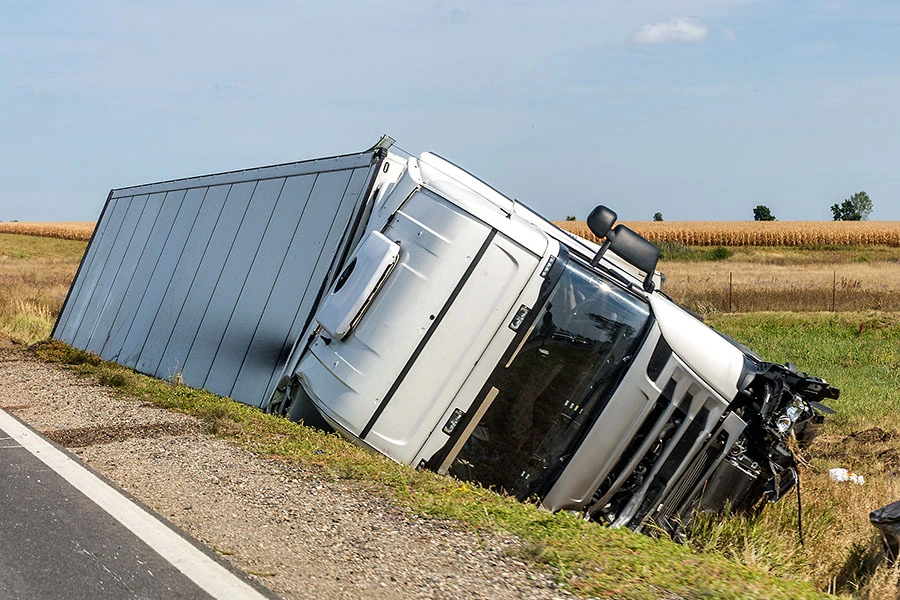 A white semi-truck lying on its side in a grassy ditch along the edge of a road.