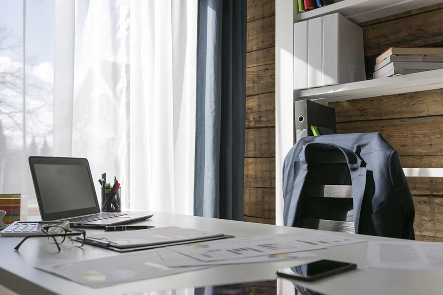 An empty desk setup with an open laptop, documents, glasses, and a jacket draped over a chair next to a window.