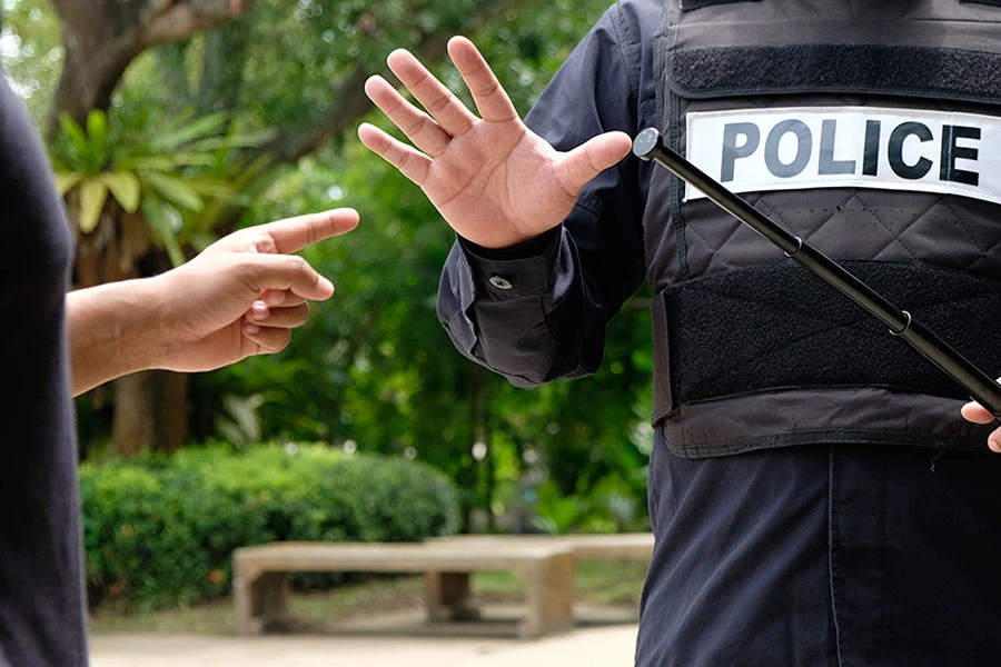 A police officer holding a baton and raising an open hand to signal "stop" toward a pointing civilian outdoors.