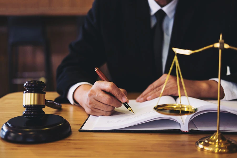 A person in a suit writing in a book at a desk with a gavel and scale of justice.
