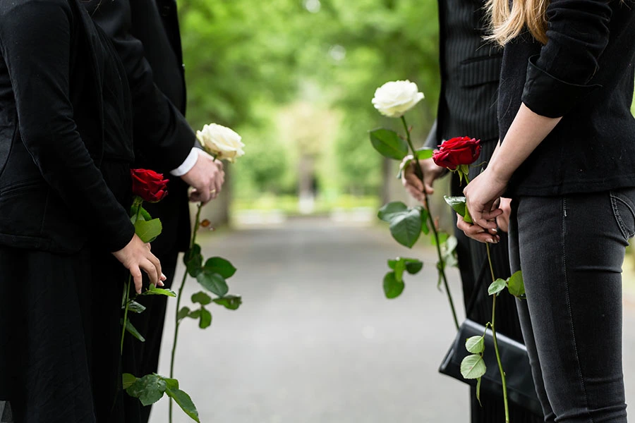 Mourners dressed in black holding single red and white roses standing along an outdoor path.
