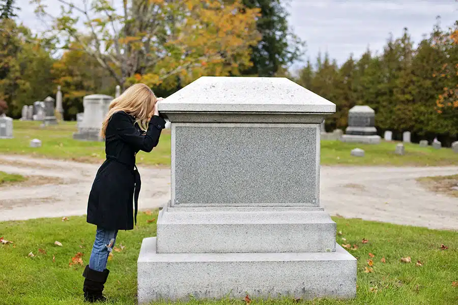 A woman leaning her face in her hands against a large stone tomb monument in a cemetery.
