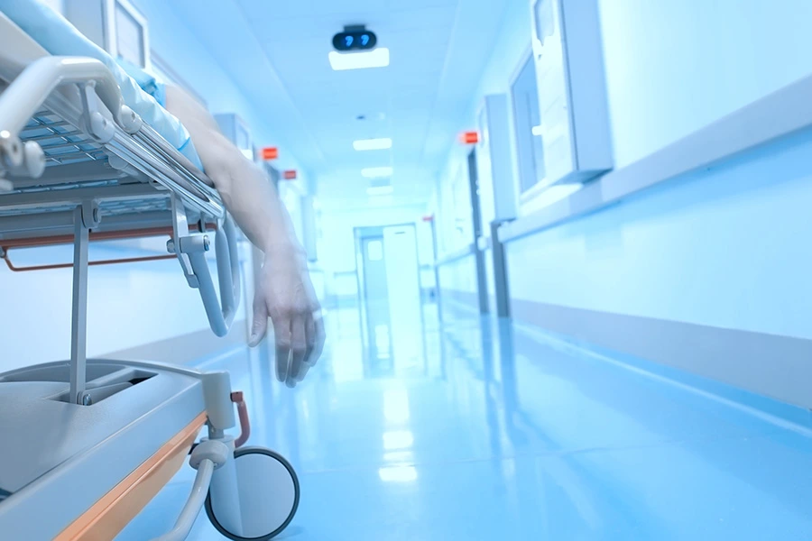 A patient's arm hangs limply from a gurney in a brightly lit, blue hospital hallway.