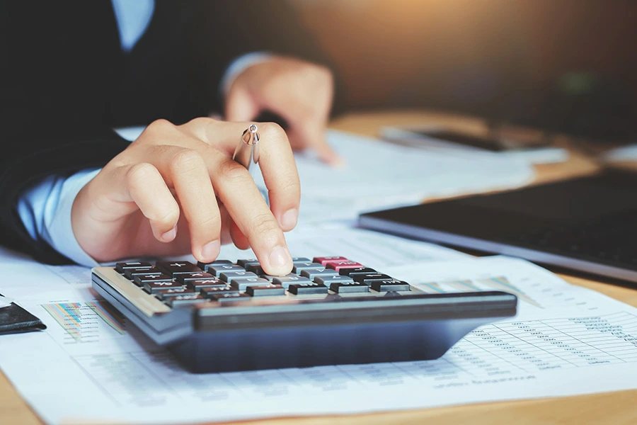 A close-up of a person holding a pen while using a calculator over financial documents on a desk
