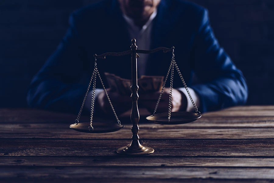 A bronze scale of justice on a wooden desk with a man in a blue suit holding money blurred in the background.