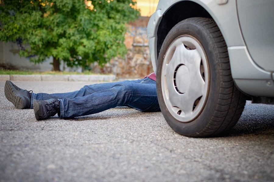 A person in jeans lying on the asphalt underneath the front wheel of a silver car following an accident.